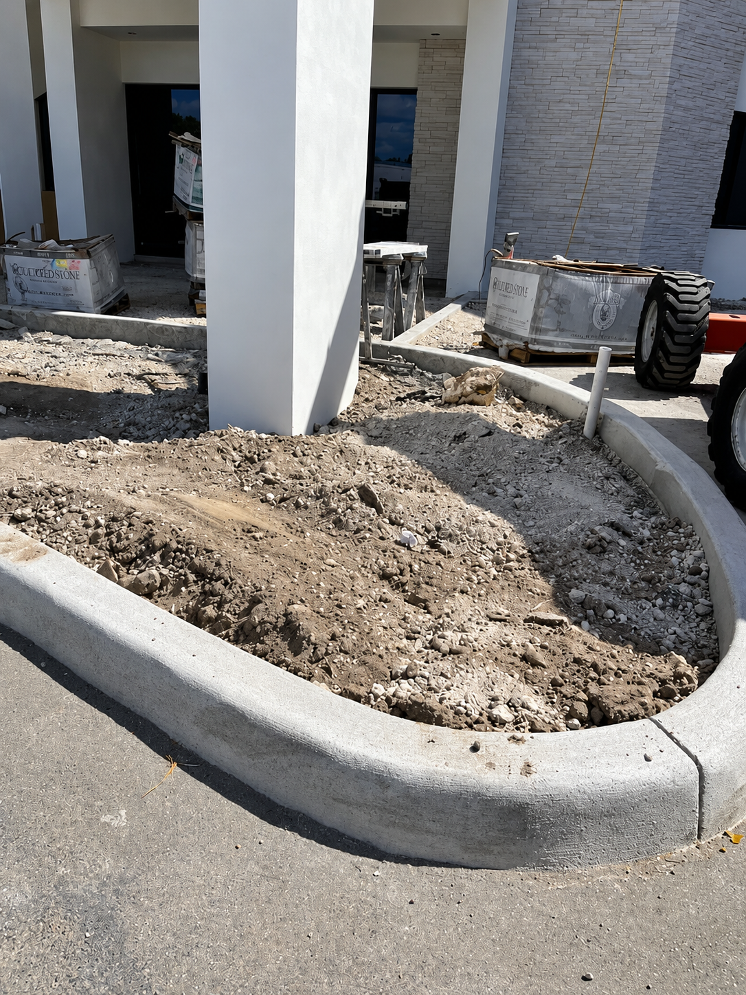 Landscaped curb with bare soil around a white column beside a brick building and sidewalk