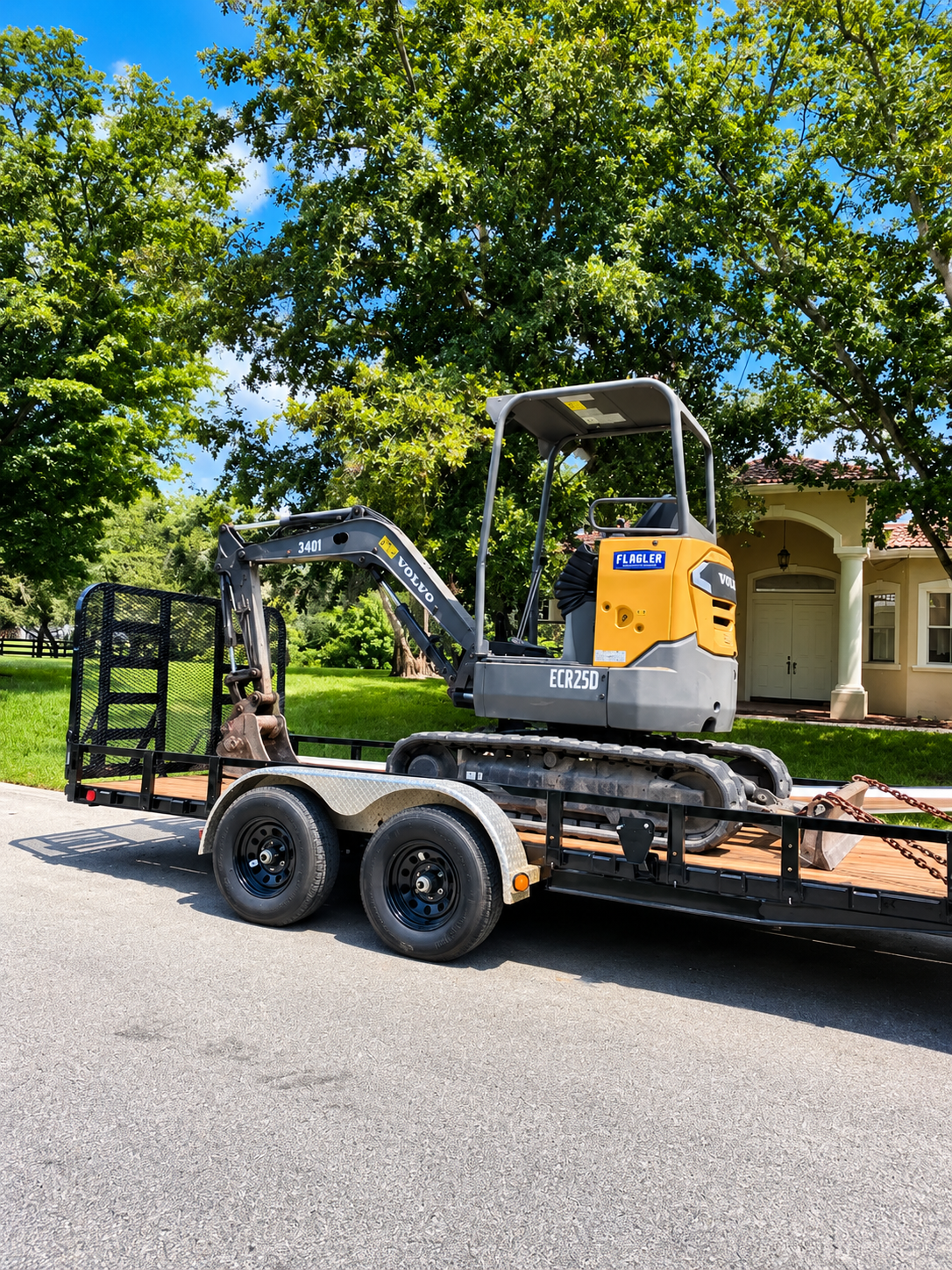 Small yellow excavator on a black flatbed trailer parked beside a house and trees