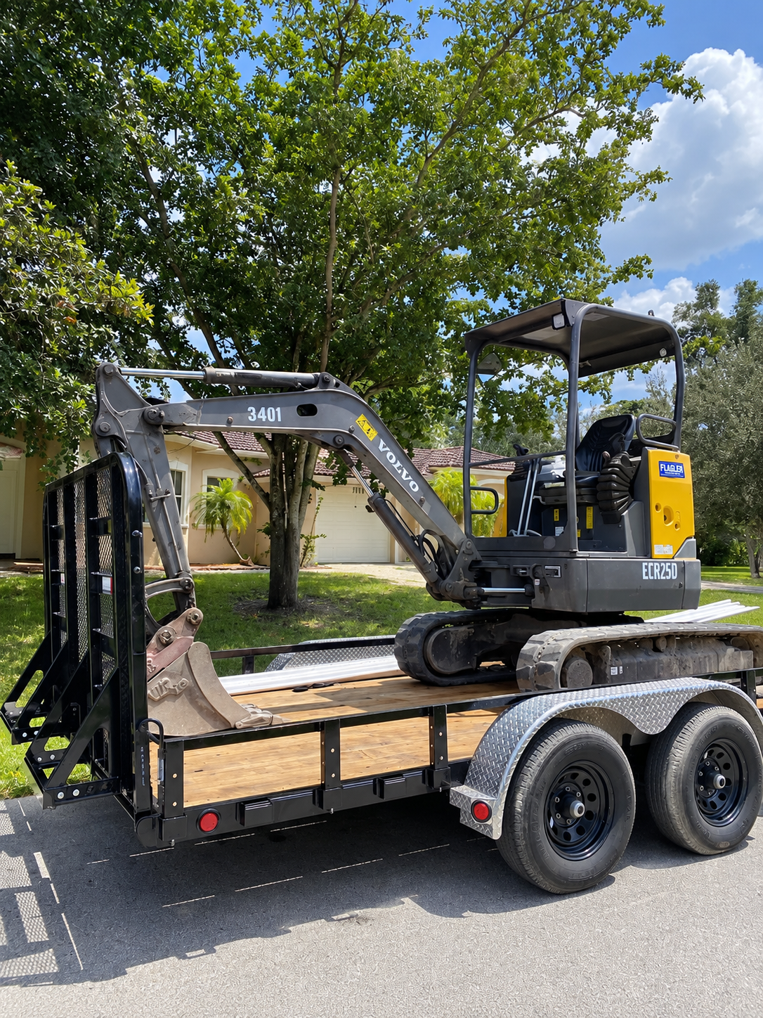 Mini excavator on a flatbed trailer under a tree, parked on a residential street.