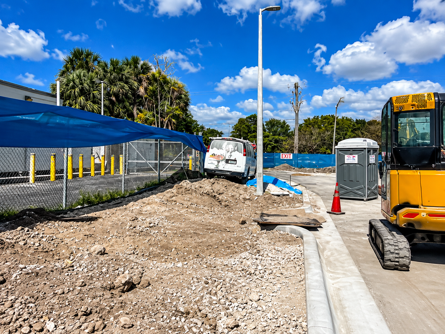Construction site with excavator, dirt piles, orange cones, and a white van by a blue fence under a bright sky