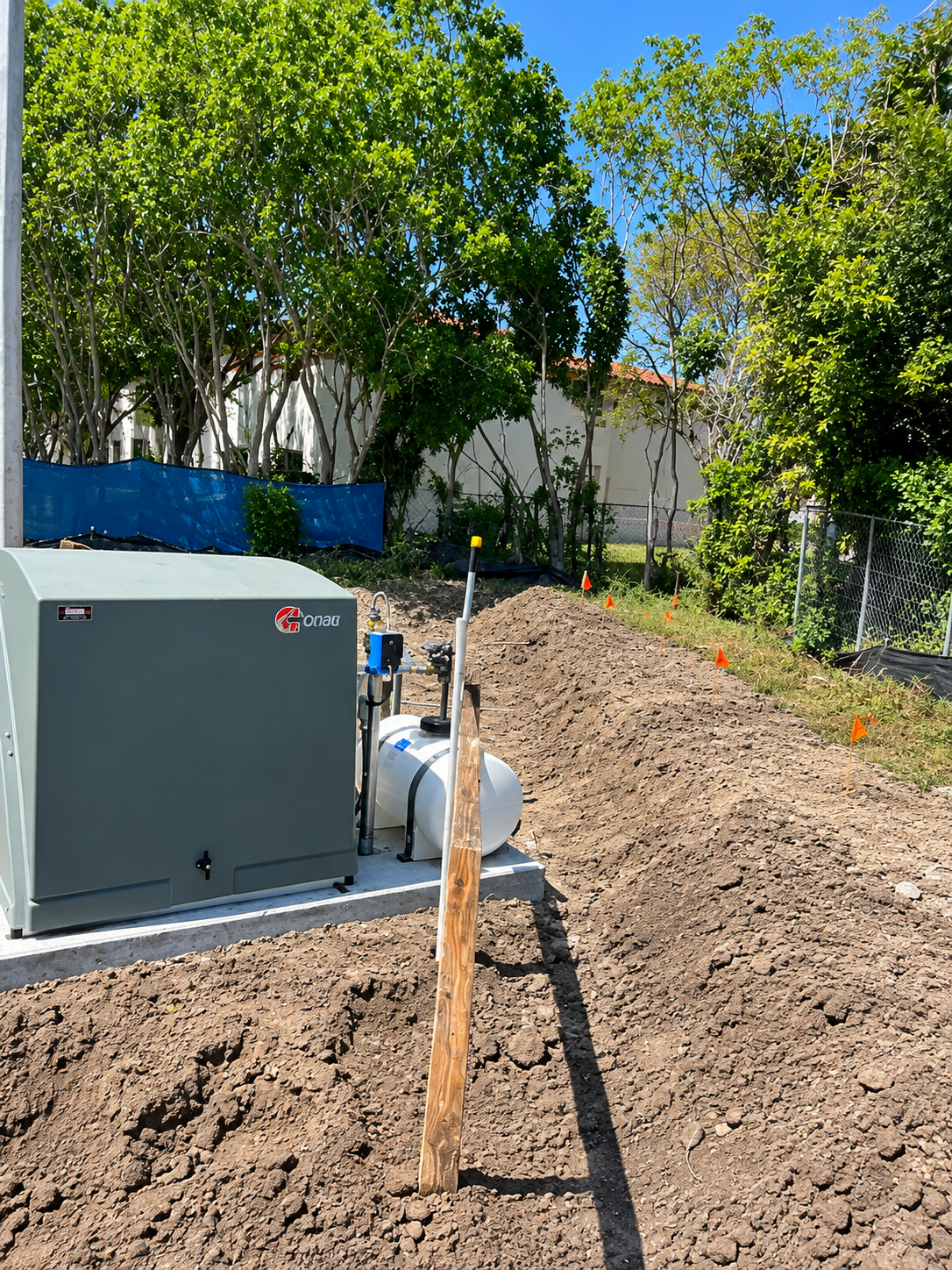 Outdoor utility area with gray equipment box, pipes, dirt mound, and newly planted tree under bright sun