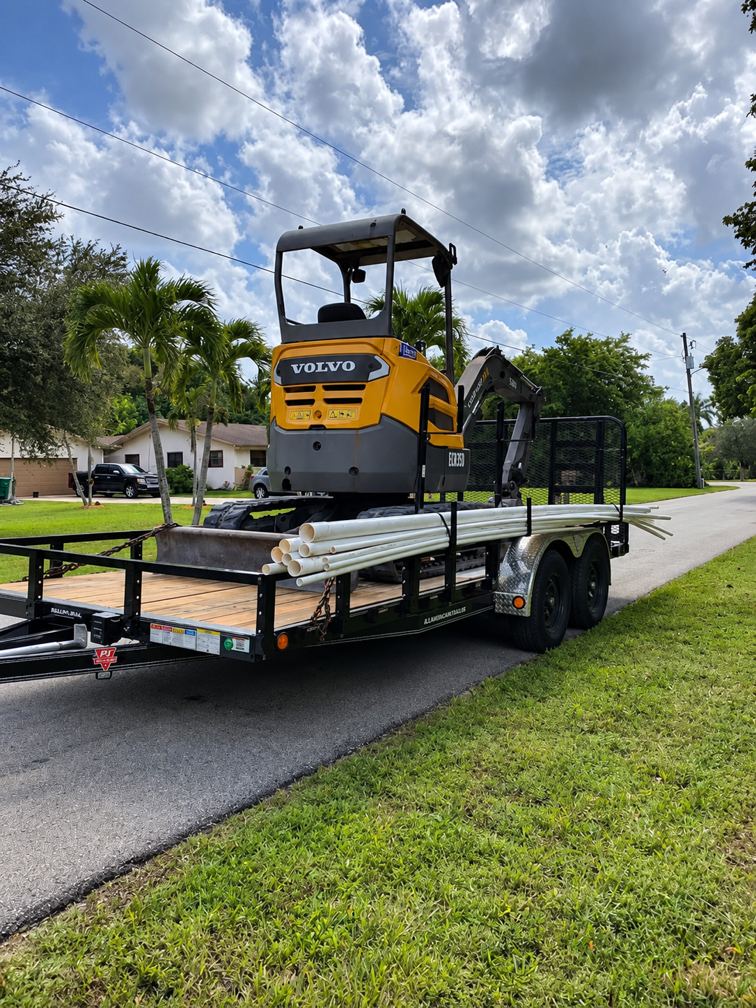 Yellow compact excavator on a flatbed trailer parked beside a grassy roadside under a cloudy sky