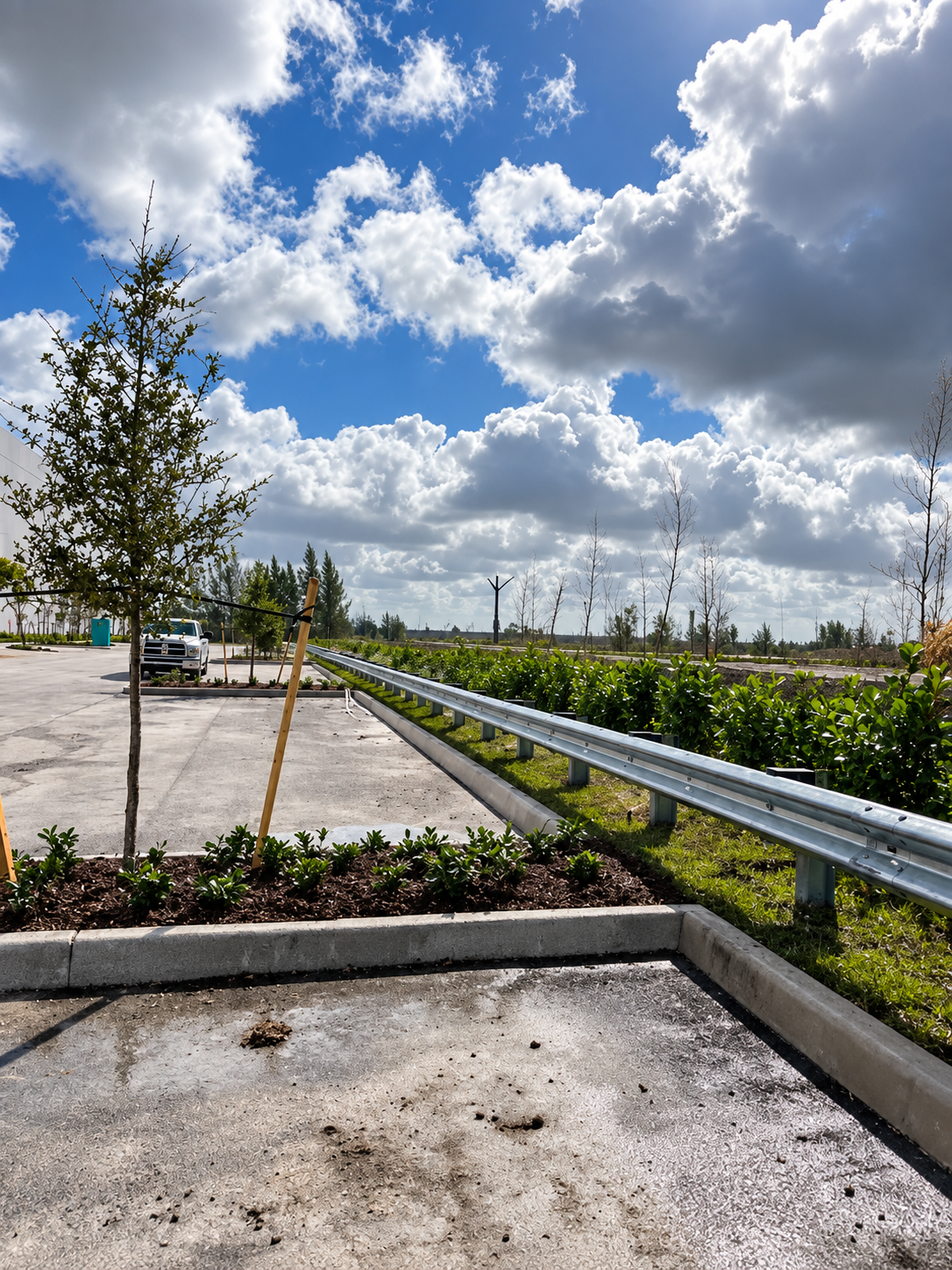 Roadside parking lot with young trees, guardrail, and cloudy blue sky over landscaped greenery.