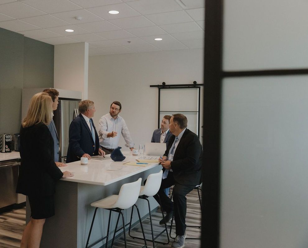 A group of people are sitting at a table in a kitchen talking to each other.