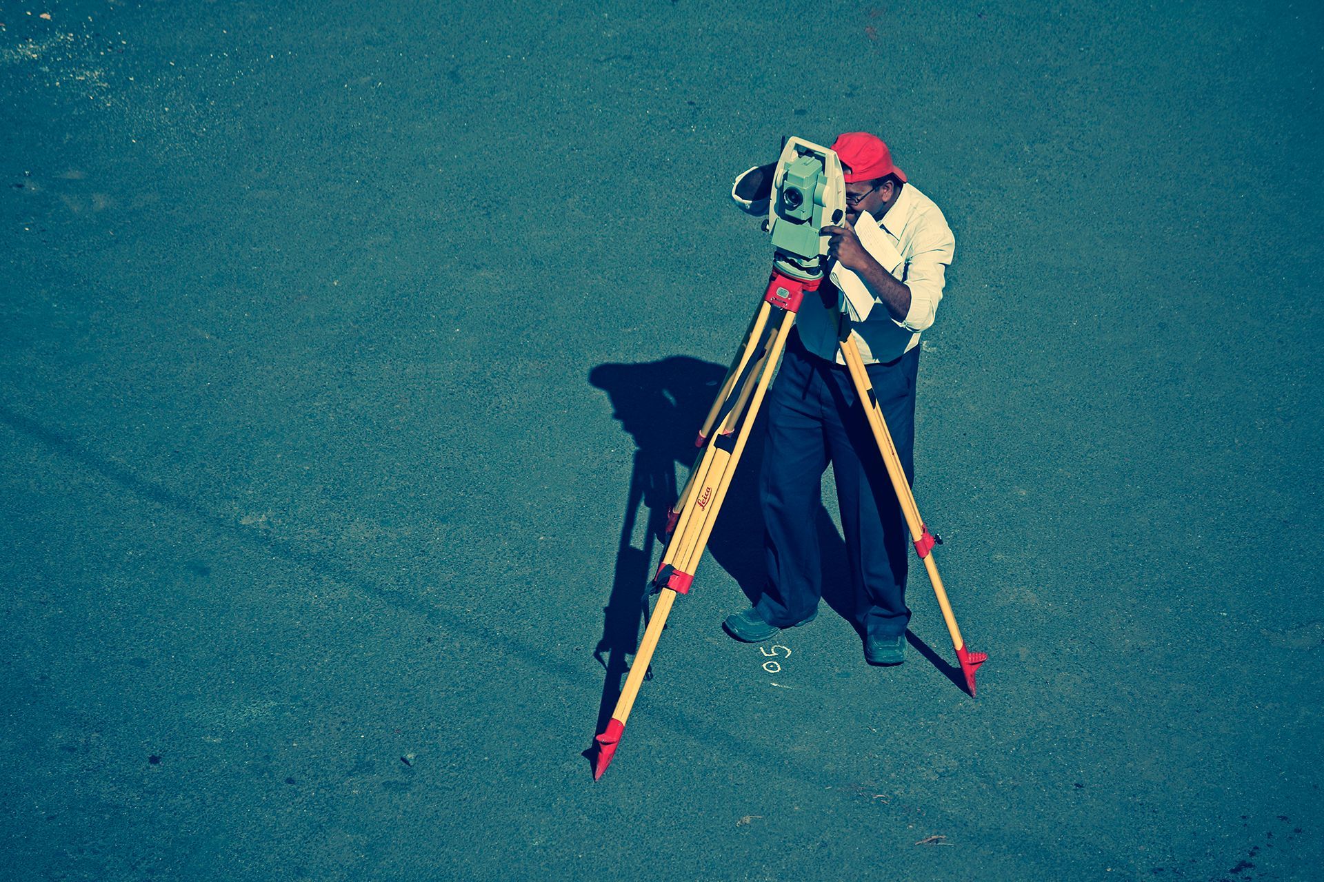 Surveyor in a red cap using a theodolite on a tripod in a paved area.