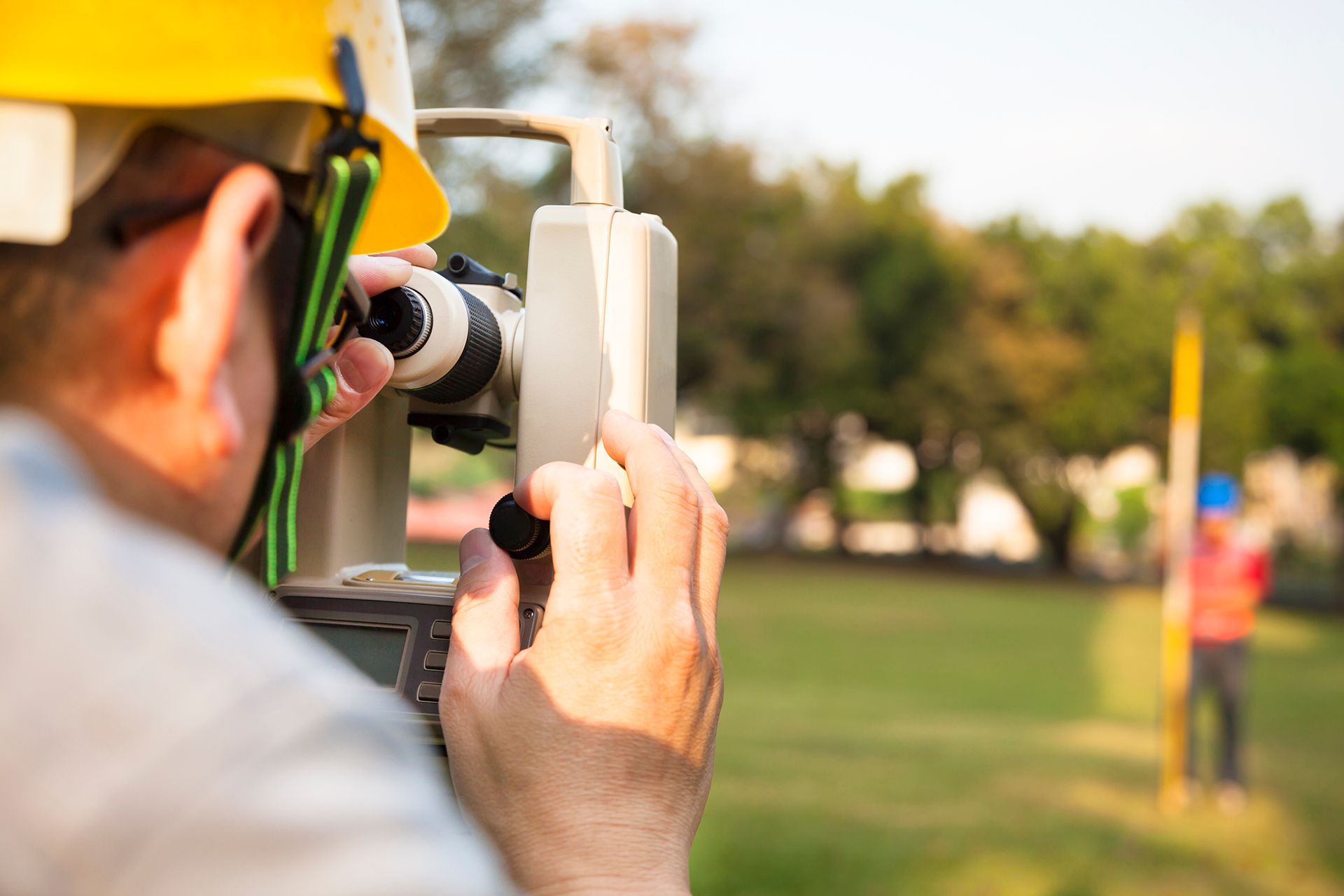 Surveyor looking through instrument, focusing on a staff held by a colleague in a grassy area.