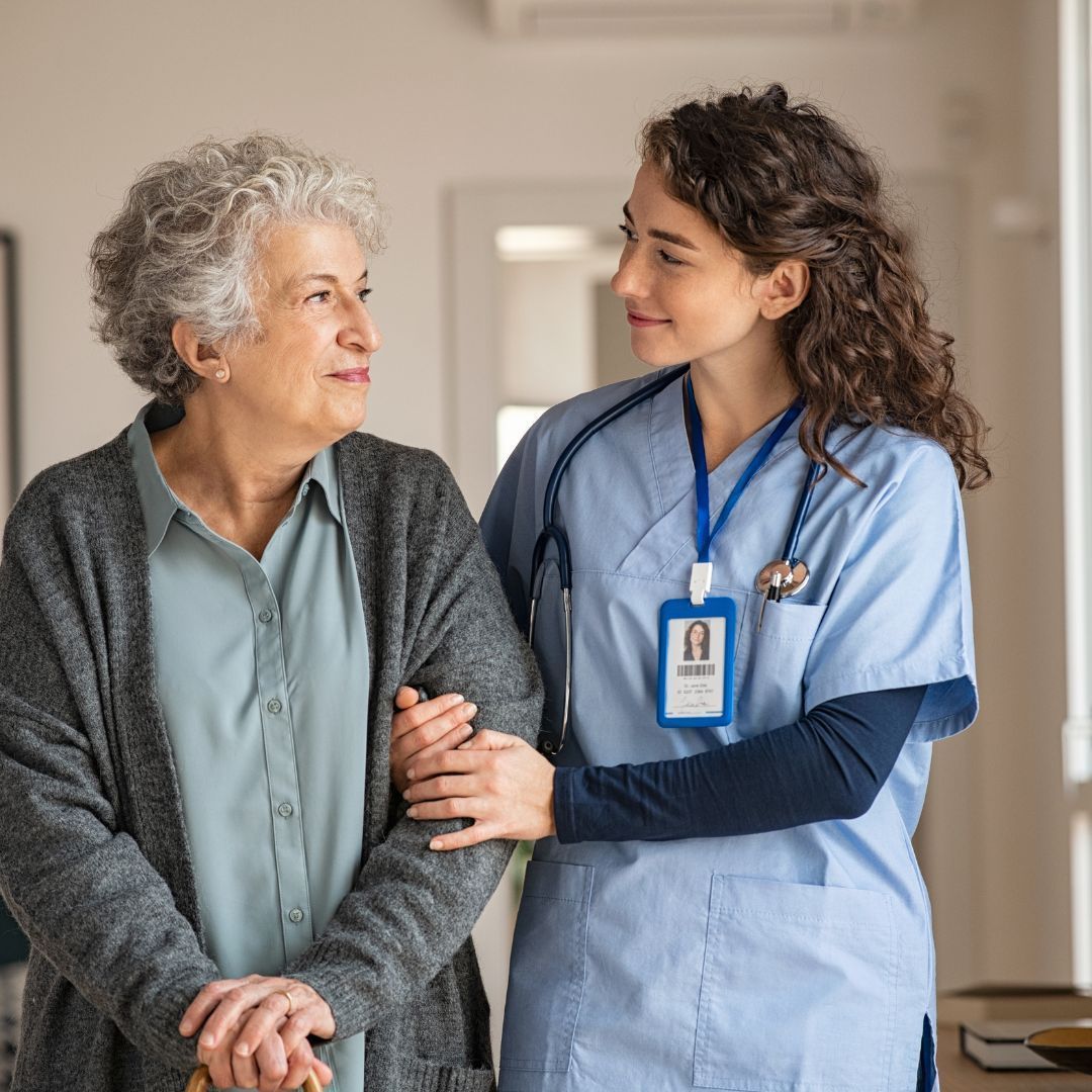 Nurse assisting an older person with a cane to walk indoors. They are looking at each other, smiling.