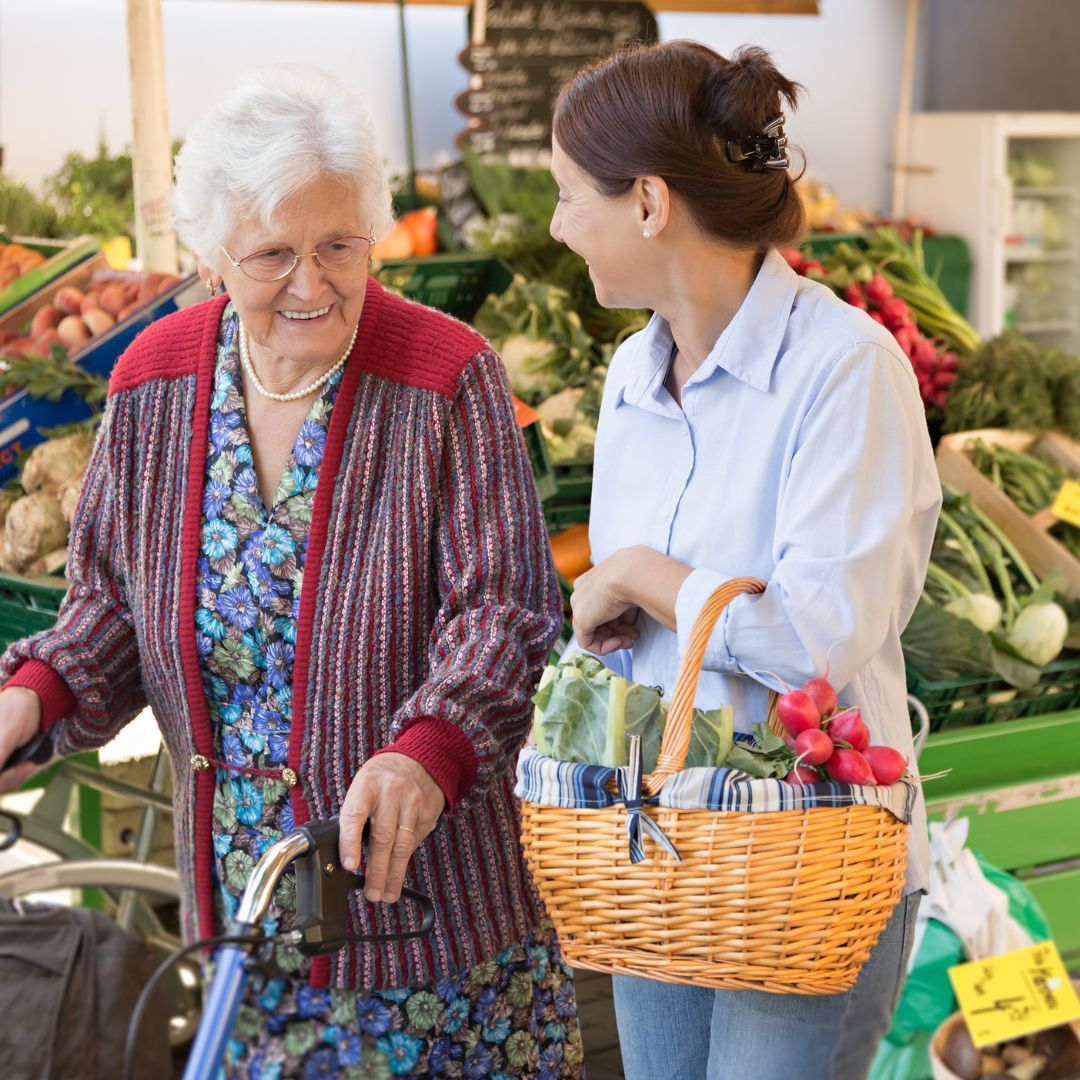 Elderly woman using a walker and younger woman shopping at a market, holding basket with produce.