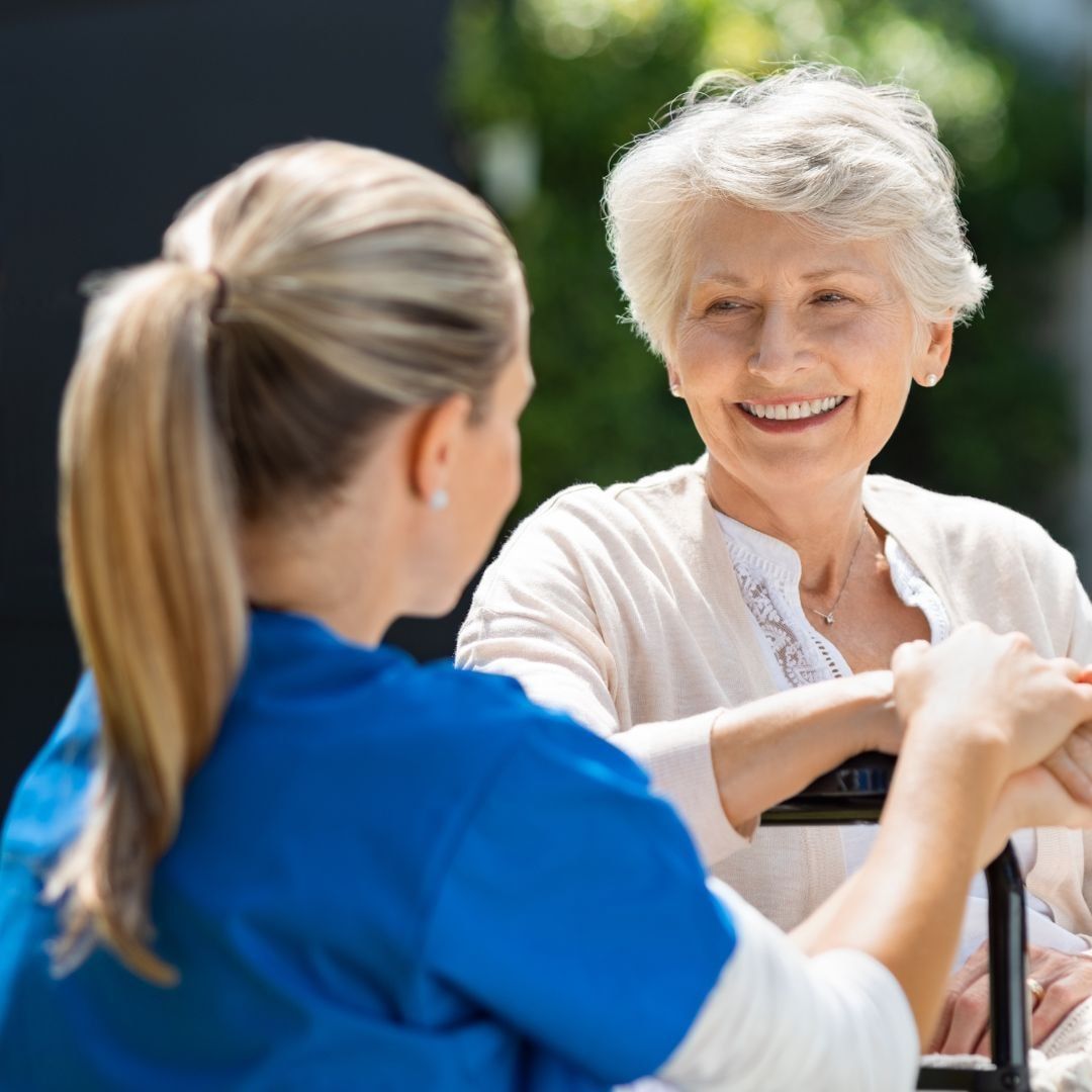 Woman in wheelchair smiles at caregiver in blue scrubs outdoors.