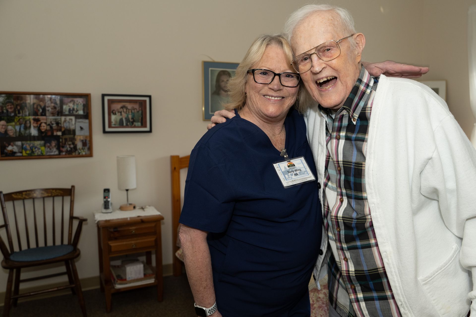 Woman in blue scrubs embraces elderly man; both smile. They are indoors in a room with photos and furniture.