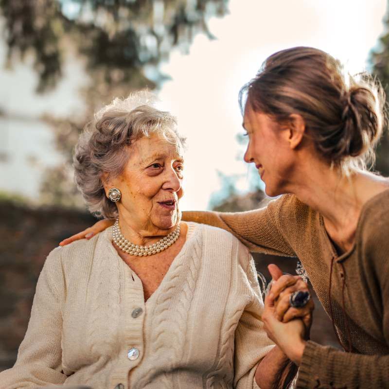 Woman comforts an older person, outdoors. One hand on their arm, the other holding theirs.