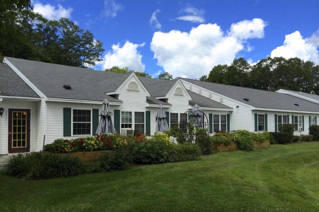 White building with green shutters, flower beds, and a grassy lawn under a partly cloudy sky.