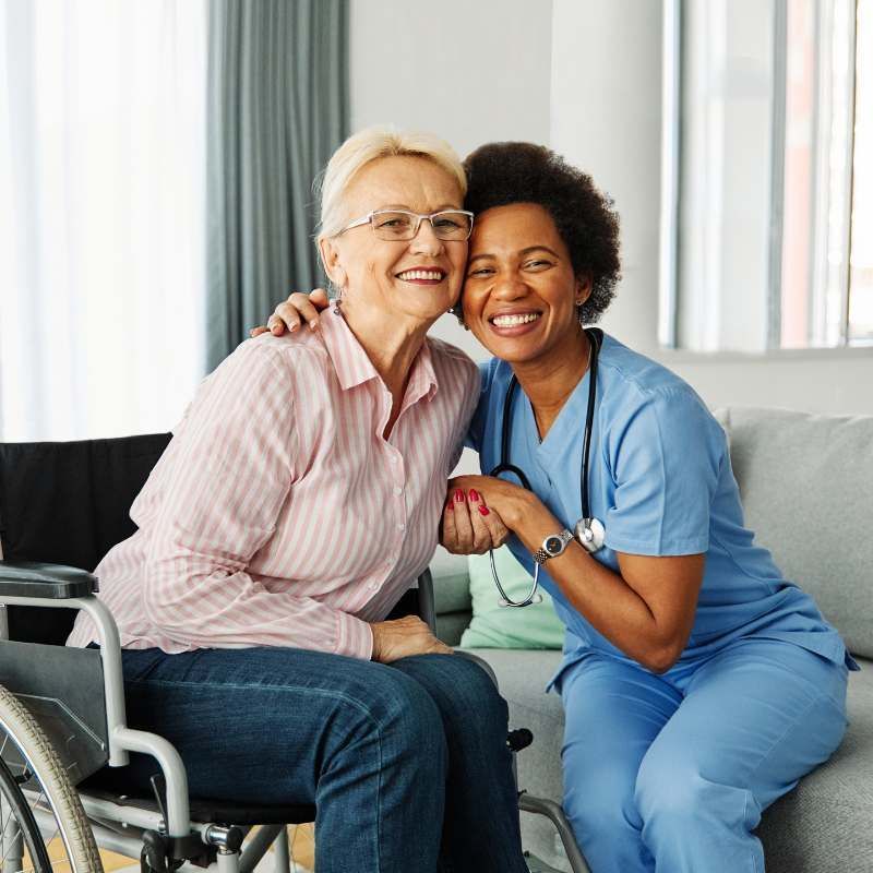 Woman in wheelchair, smiling, embraced by caregiver. Both looking at camera. Indoors.