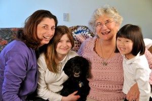 Family of four smiling with a black dog, seated indoors. A woman wears purple, another a pink sweater. Two girls.