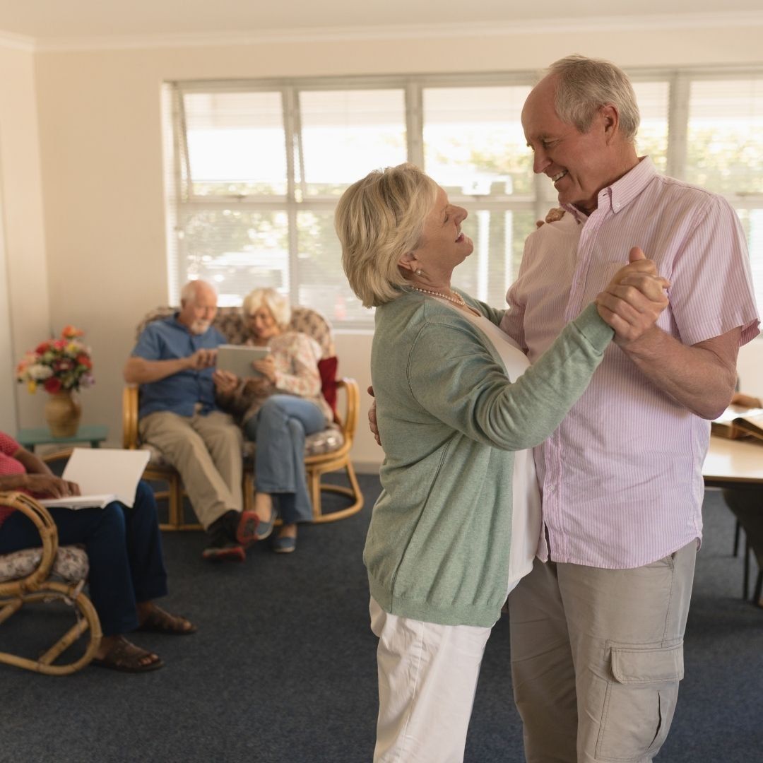 Couple dancing in a room, smiling. Others seated, watching. Bright room, sunlight.