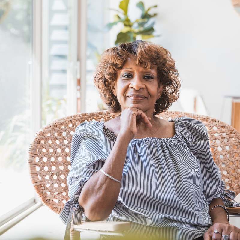 Woman sitting in a wicker chair, looking at the camera with her hand on her chin.