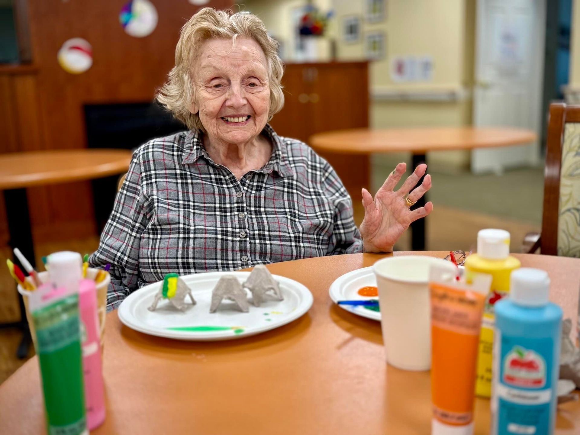Woman smiles and waves at a table with paint and egg carton art in a room.