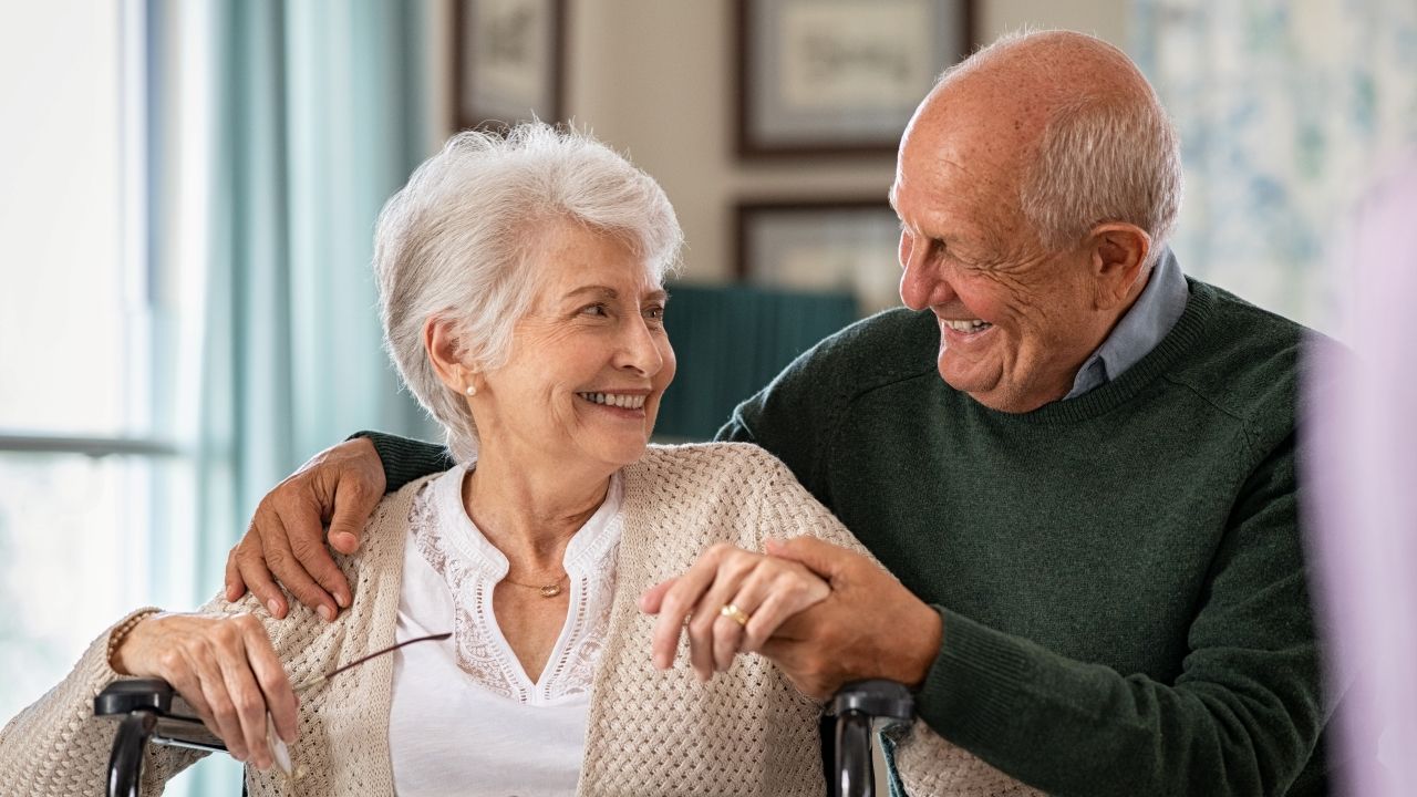 A smiling person in a knit cardigan with glasses sits in a chair while another person in a green sweater rests a hand.
