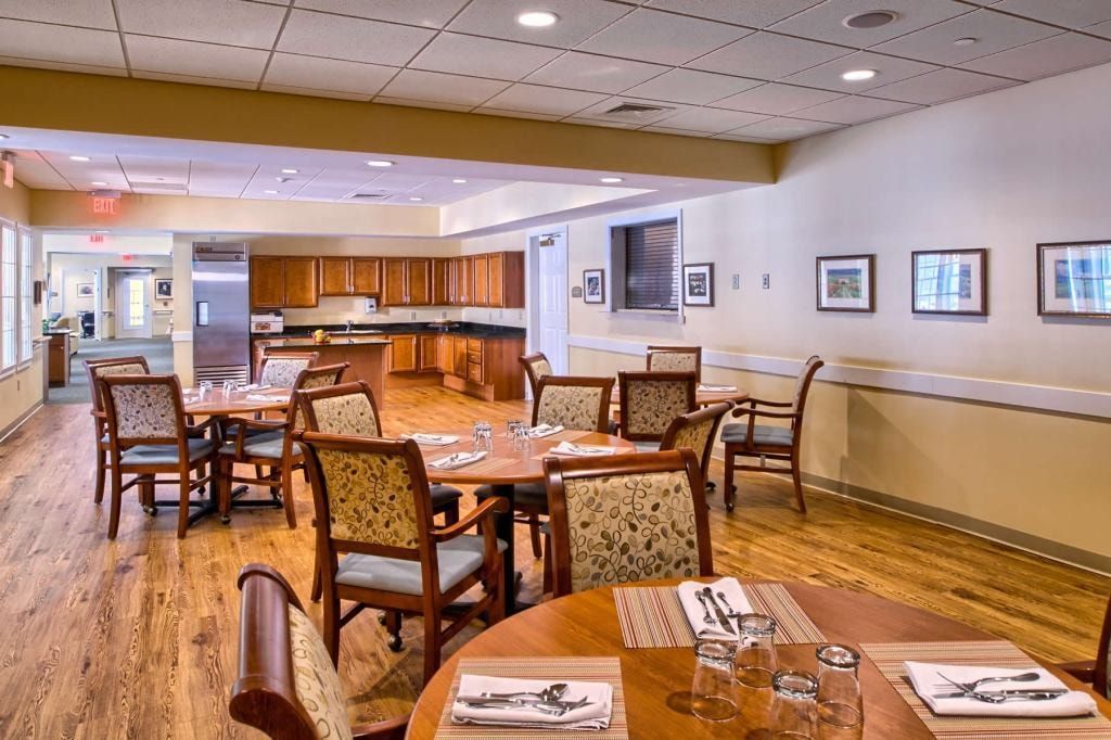 Dining room with wooden tables, chairs, and a kitchen in the background. Light-colored walls and floor.