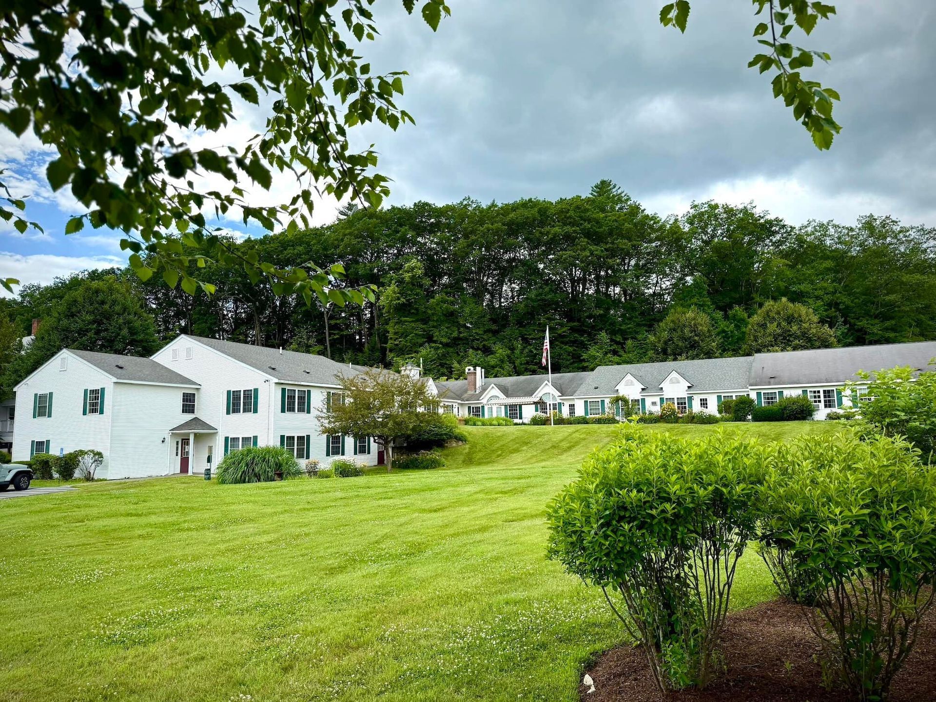 White buildings with green shutters sit on a grassy hill, surrounded by trees under a cloudy sky.