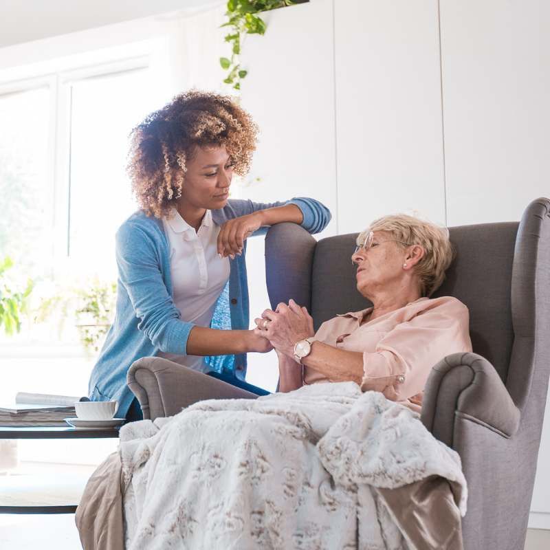 Woman comforts older person in a chair; holds hands, looks concerned. Bright interior.