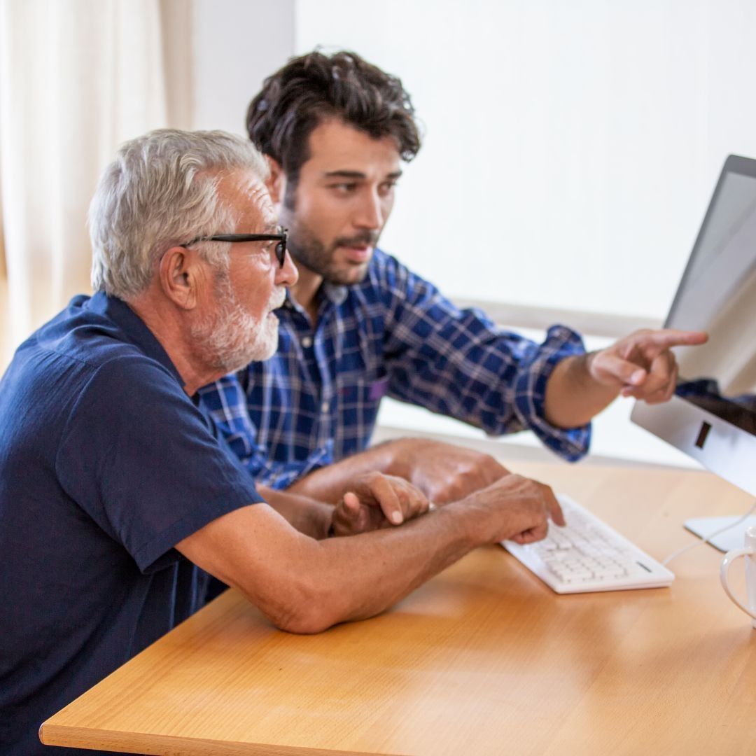 Man pointing at a computer screen, assisting an older person who types at the keyboard.
