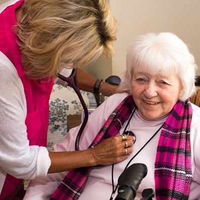 Caregiver using a stethoscope on an elderly woman sitting in a chair. Woman smiles, wears pink scarf.