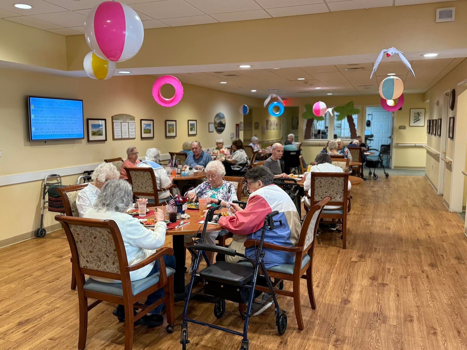 Seniors playing games at tables in a decorated activity room. Beach balls and floats hang from the ceiling.