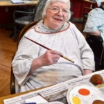 Woman in painting smock holds a paintbrush, smiling at a table with paints and supplies.