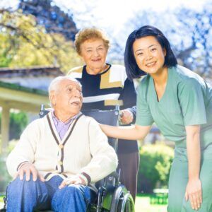 Woman in scrubs assisting an elderly man in a wheelchair and an older woman outside.