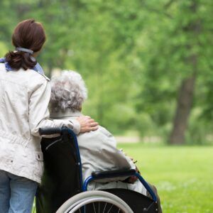Woman in a white jacket assisting a person in a wheelchair outdoors, arm around their shoulder.