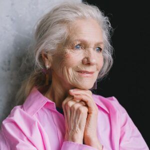 Woman with silver hair, wearing a pink shirt, hands clasped, looking thoughtful.