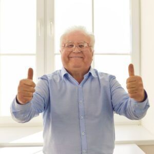 Man in a light blue shirt giving double thumbs up, smiling indoors.