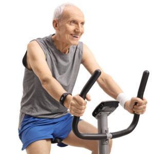 Older person exercising on a stationary bike, smiling. Wearing gray tank top, blue shorts, and a fitness tracker.