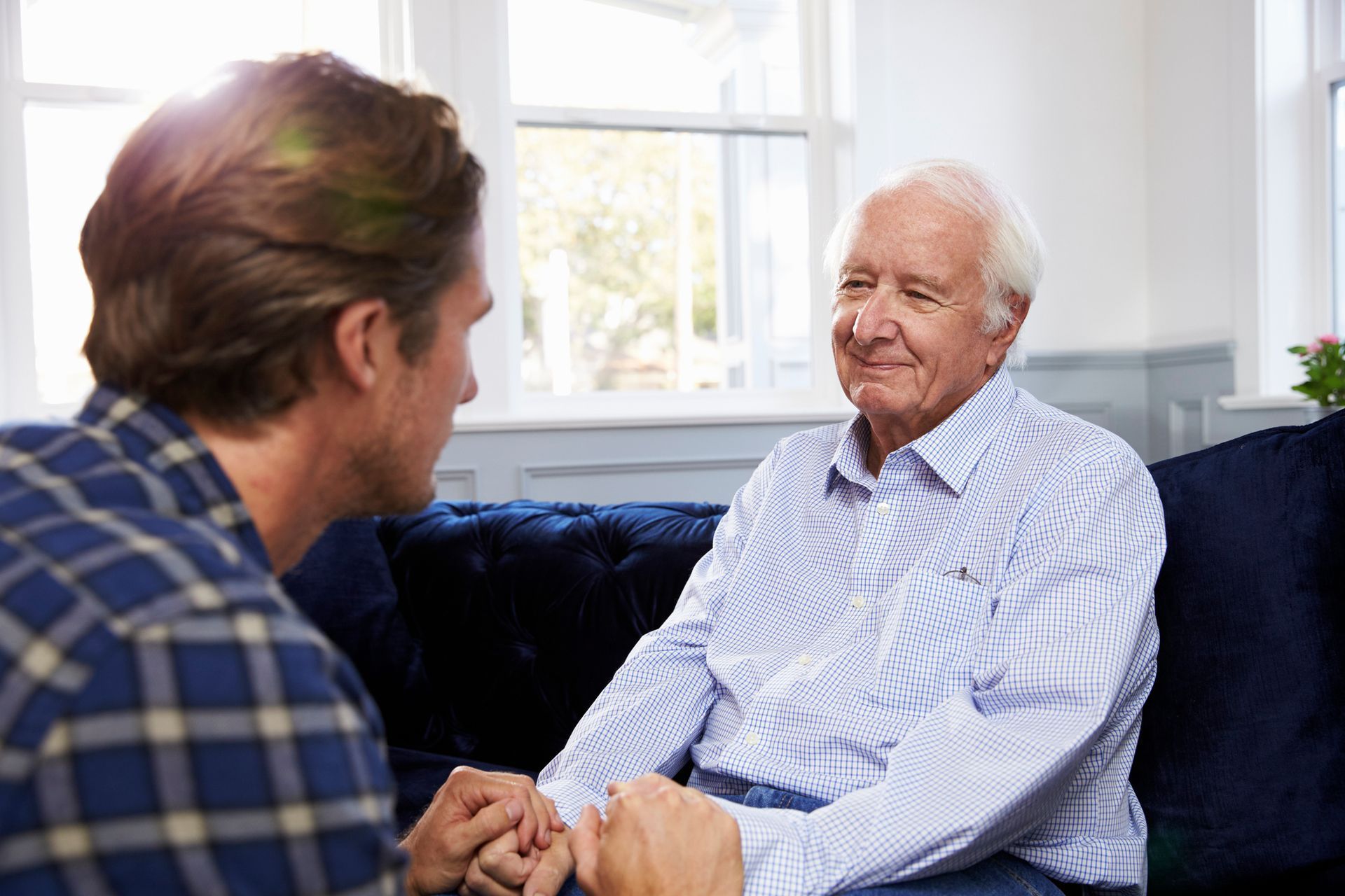 Adult son holding hands with his elderly father while talking on a couch