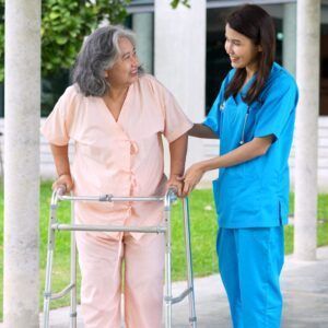 Nurse assists patient using a walker outdoors; the patient smiles, wearing pink.