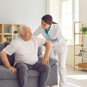 Woman in uniform assists an older adult man sitting on a sofa. Both are smiling indoors.