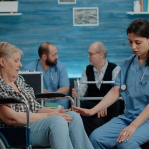 Elderly woman in wheelchair with nurse, two other people in background. Blue tones.