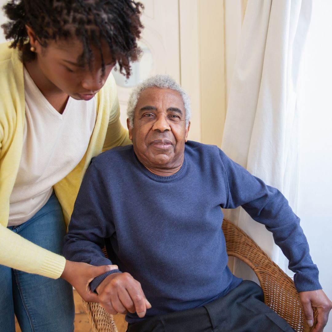 A young person helps an older person seated in a wicker chair, indoors.