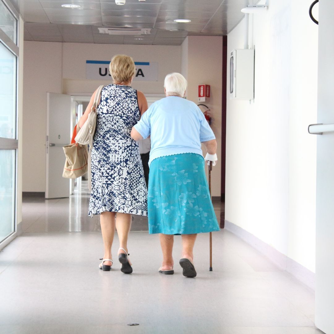 Two women walking down a hospital hallway; one using a cane, one carrying a bag.