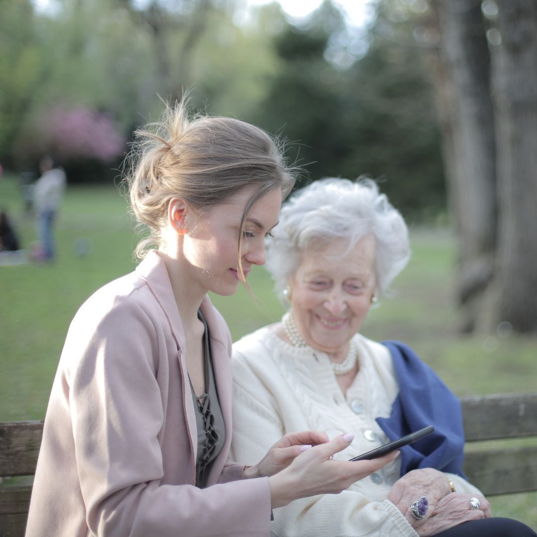 Young woman showing elderly woman something on a phone. They're sitting on a park bench, looking at the screen.