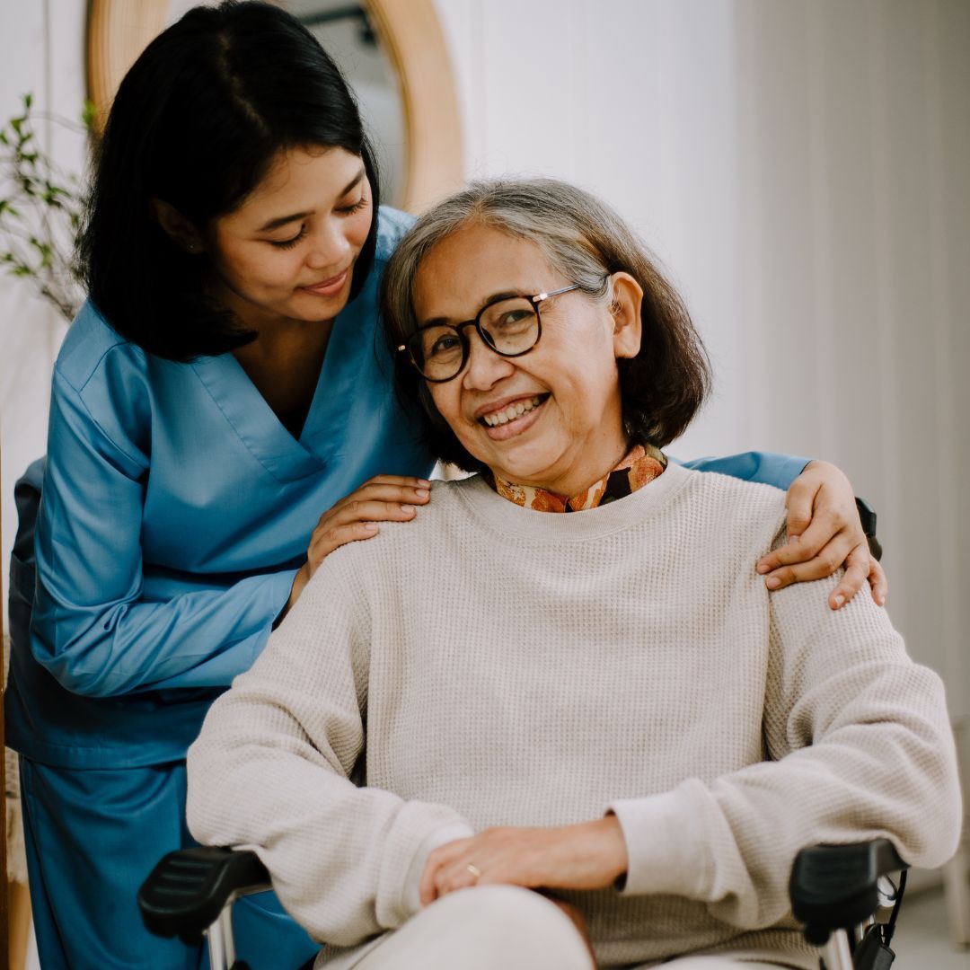 Caregiver with arms around senior in wheelchair, smiling. Blue scrubs, neutral sweater. Indoors.