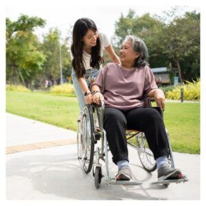 Young person assisting older person in a wheelchair outdoors; park setting; smiles.