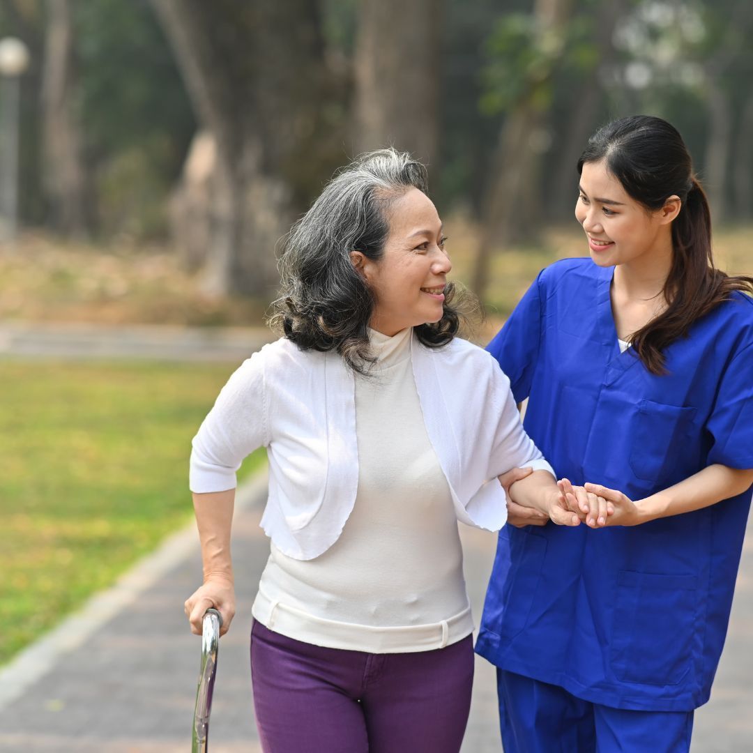 Elderly woman walks outdoors with caregiver, holding cane, smiling. Park setting, blue scrubs.