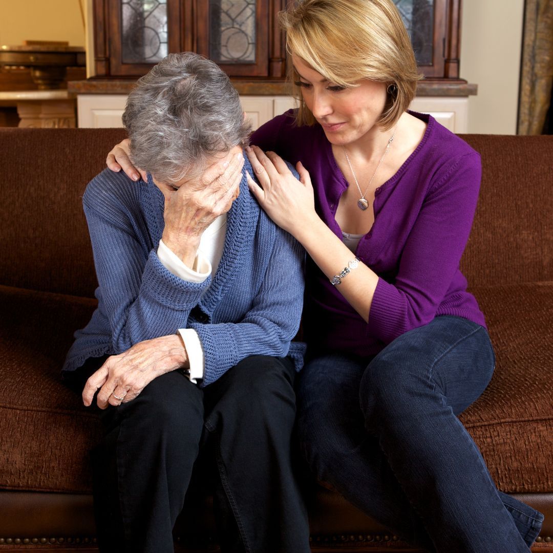 Woman comforts elderly person on a brown couch. Elderly person cries, covering face. Woman has arm on shoulder.