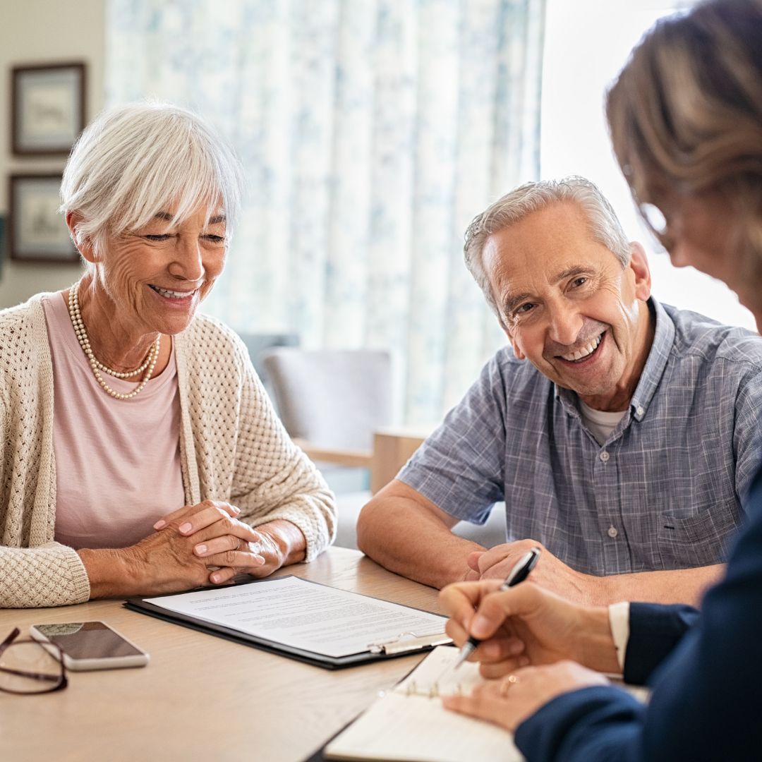 Elderly couple smiles, looks at a document, with person in suit writing at a table.