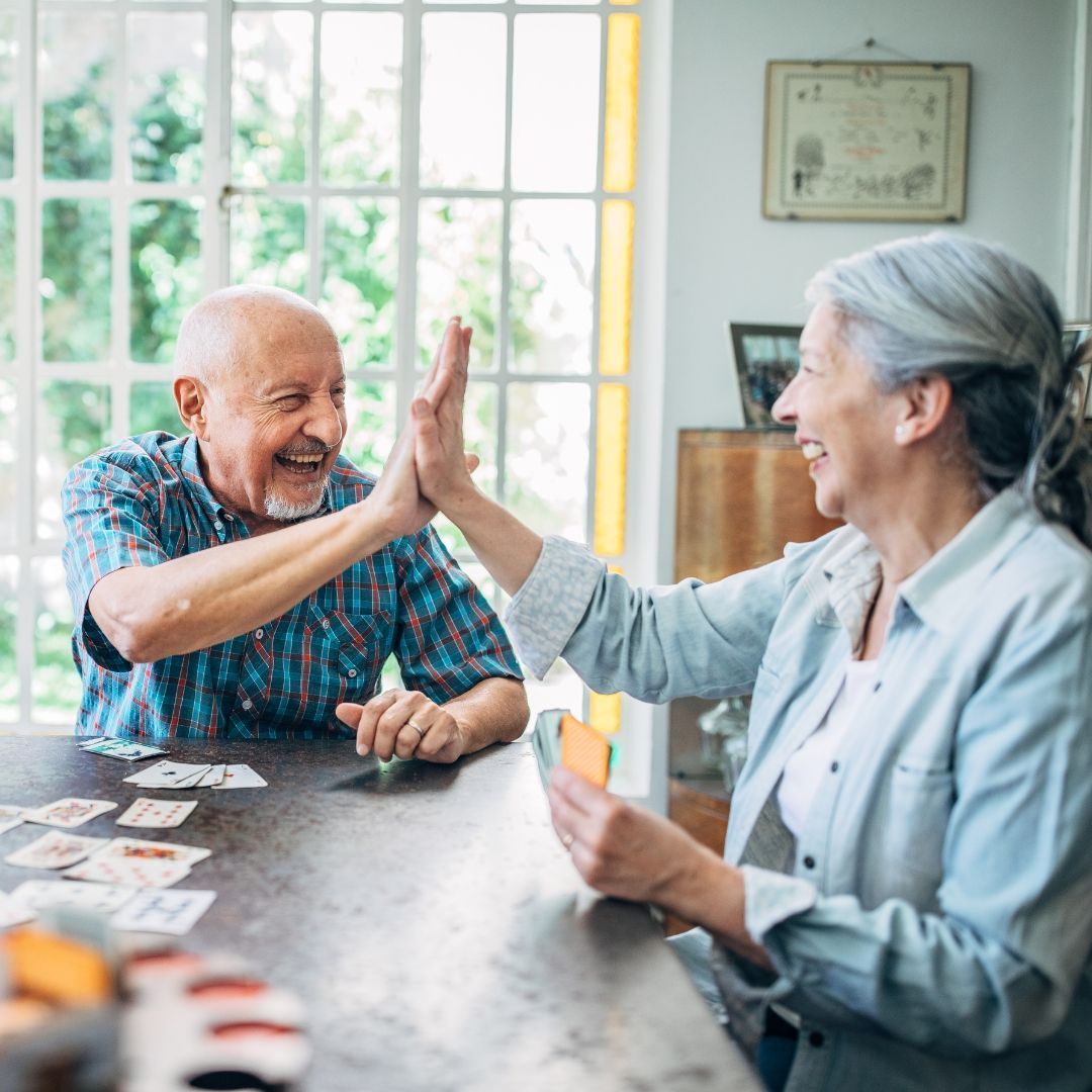 Elderly couple high-fiving over a table with playing cards in a sunlit room.