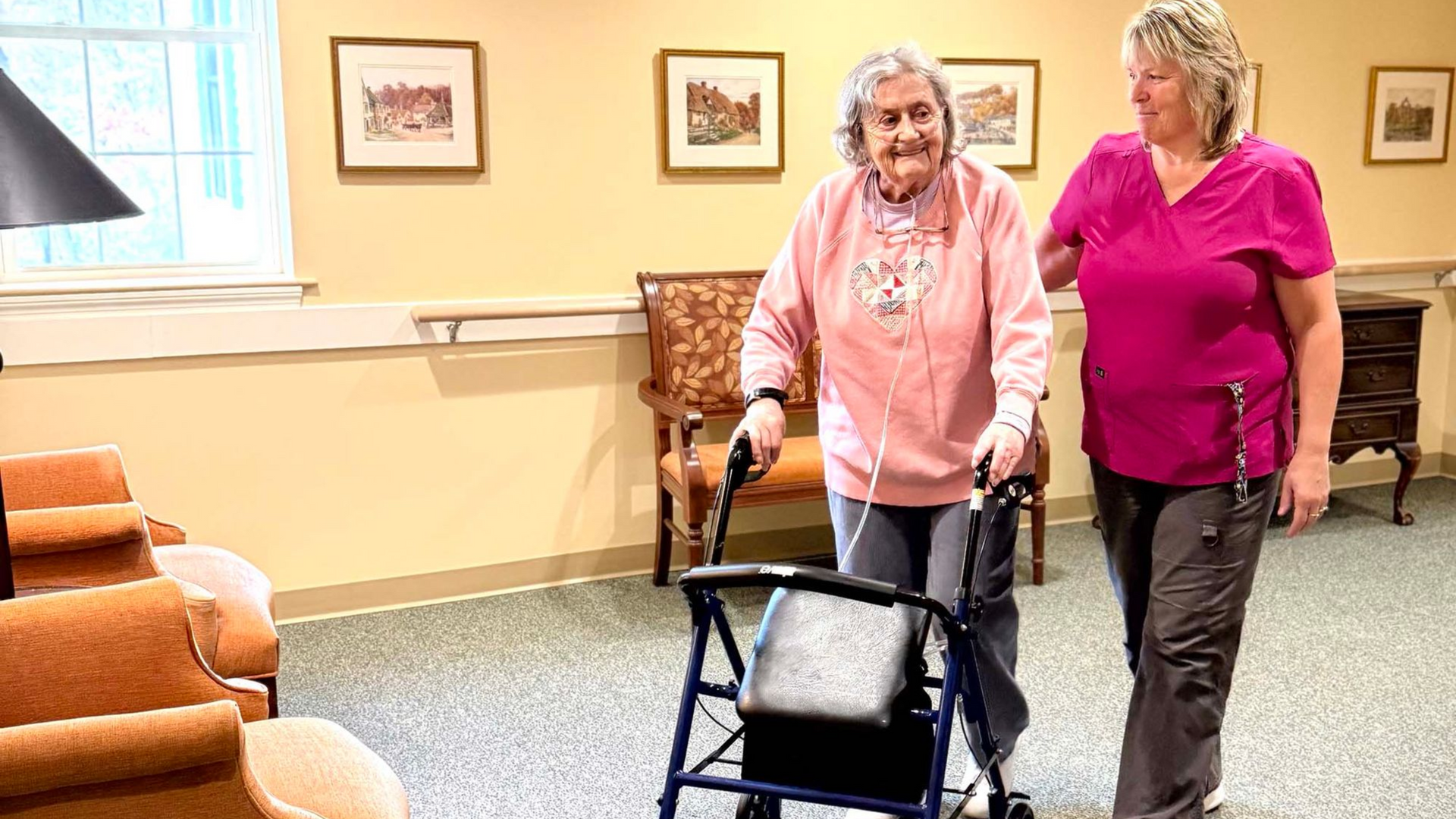 Two women walking with a rollator in a bright room with chairs and framed pictures.