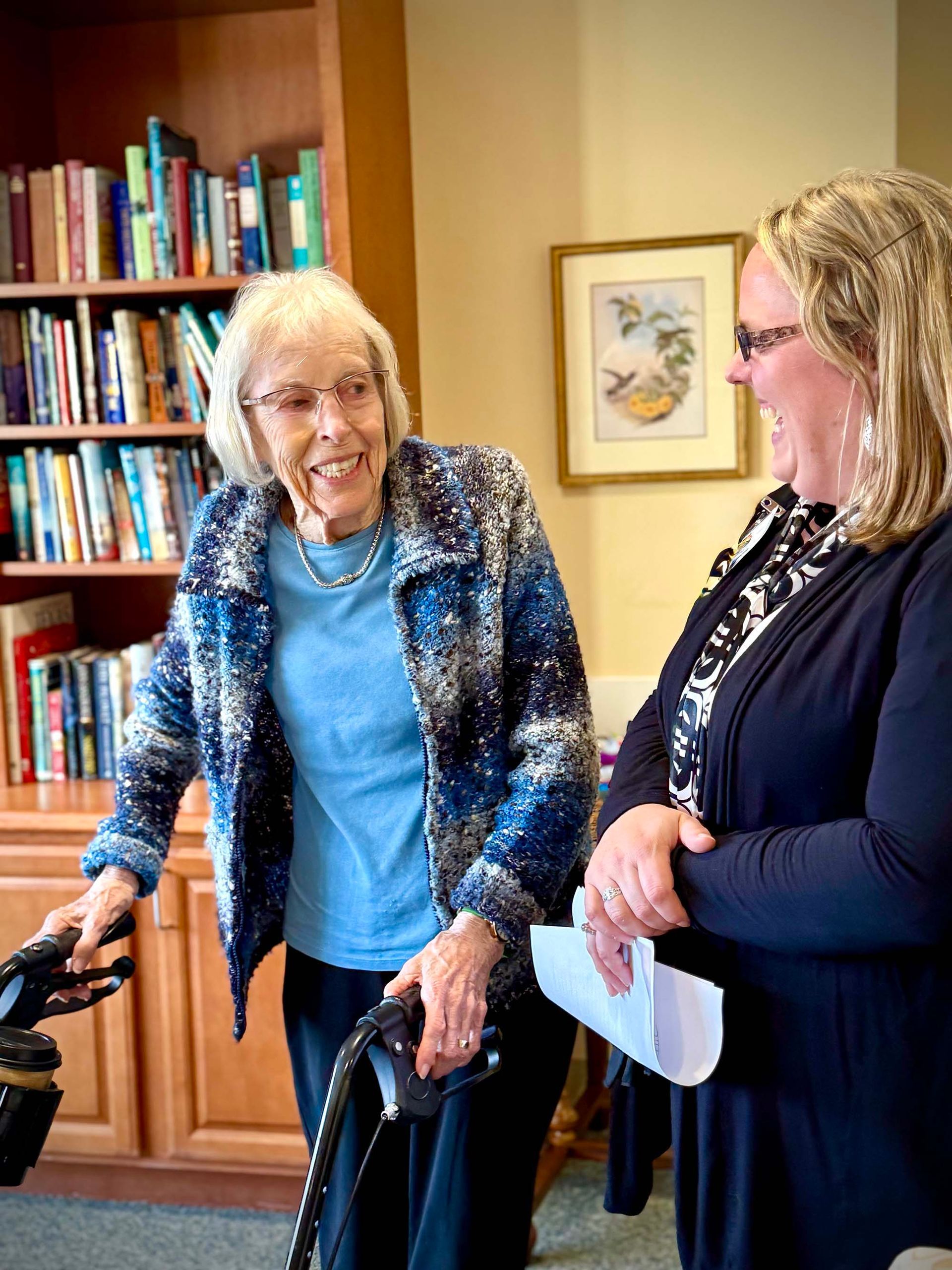 Two women smiling and talking in a cozy room, one using a walker, with bookshelves and framed art behind them.