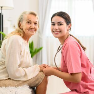 Elderly person and caregiver smiling, holding hands. Indoors, warm tones, pink scrubs.